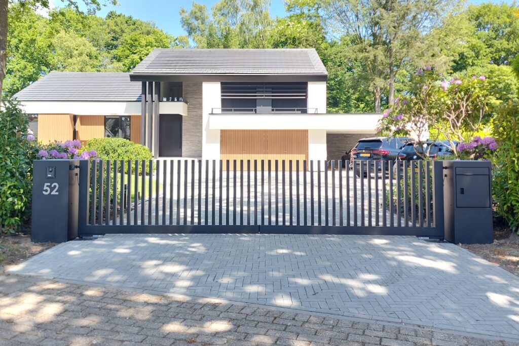 Stalen poort met verticale spijlen die aan de bovenzijde niet met elkaar verbonden. Het geeft een open karakter aan de poort. De poort heeft aan beide zijden stalen kolommen met huisnummer, intercom, brievenbus/pakketbox. Een prachtig geheel i.c.m. de woning.