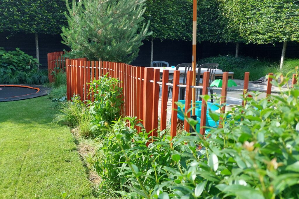 Houten hekwerk ca. 1,2 meter hoog. Houten padouk balkjes welke meanderend door de tuin, met de beplanting mee, zijn geplaatst als afscheiding van het zwembad. De tussenafstand van de balkjes is ongeveer 8 cm. De balkjes zijn verbonden met een stevige staalkabel.